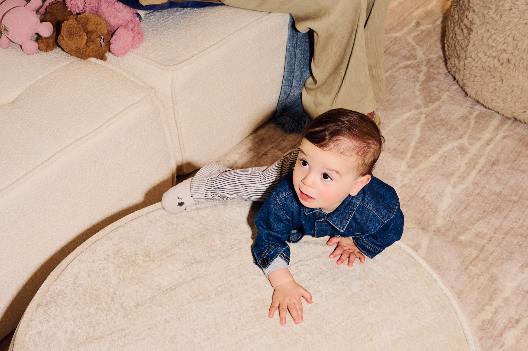 Child in a denim jacket crawling on a beige carpeted floor with toys around.