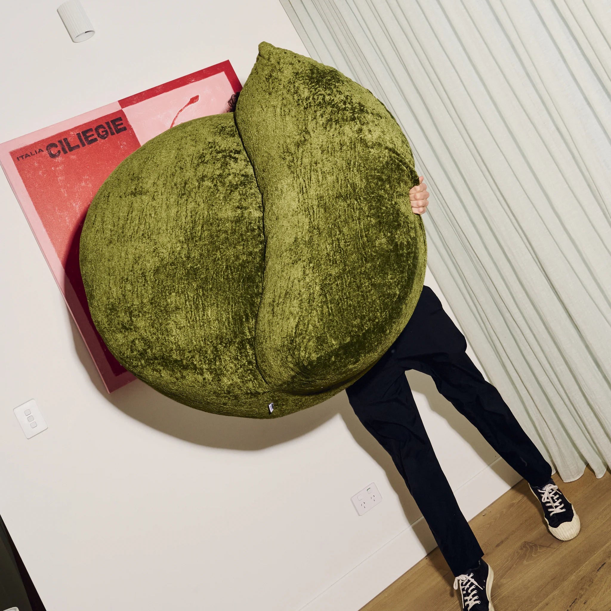 Person holding a large green cushion resembling a fruit on a white floor with a pink book.