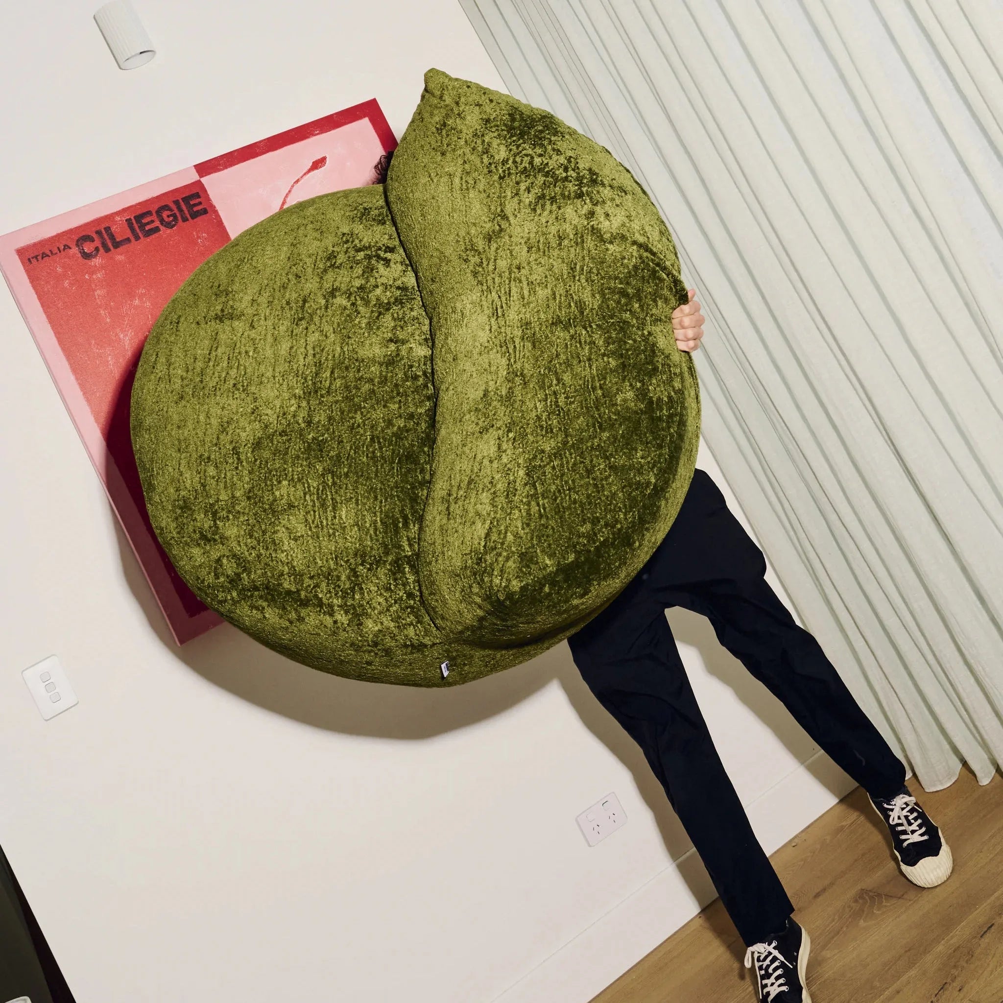 Person holding a large green cushion resembling a fruit on a white floor with a pink book.