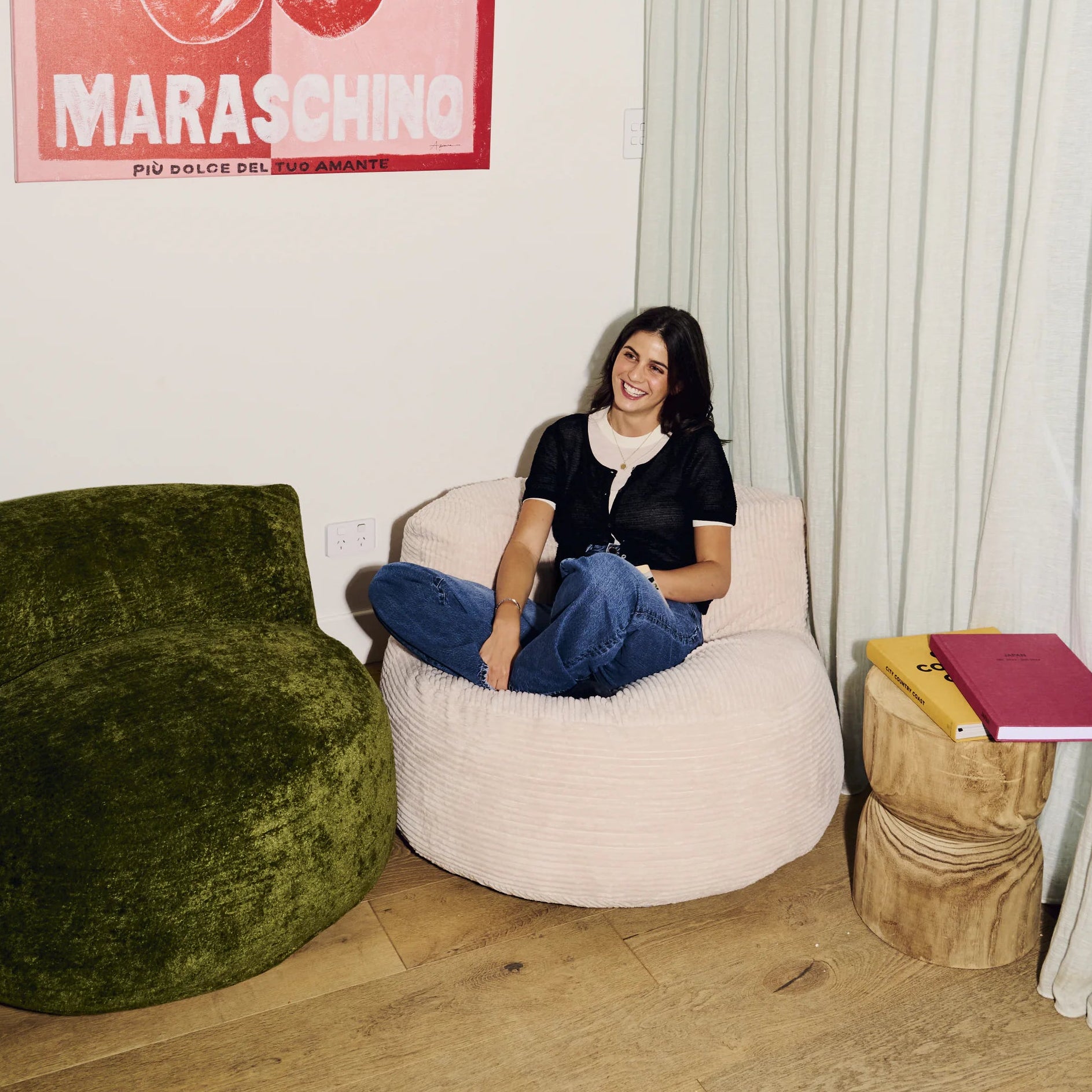 Woman sitting on a beige bean bag chair in a room with a green armchair and a pink poster on the wall.