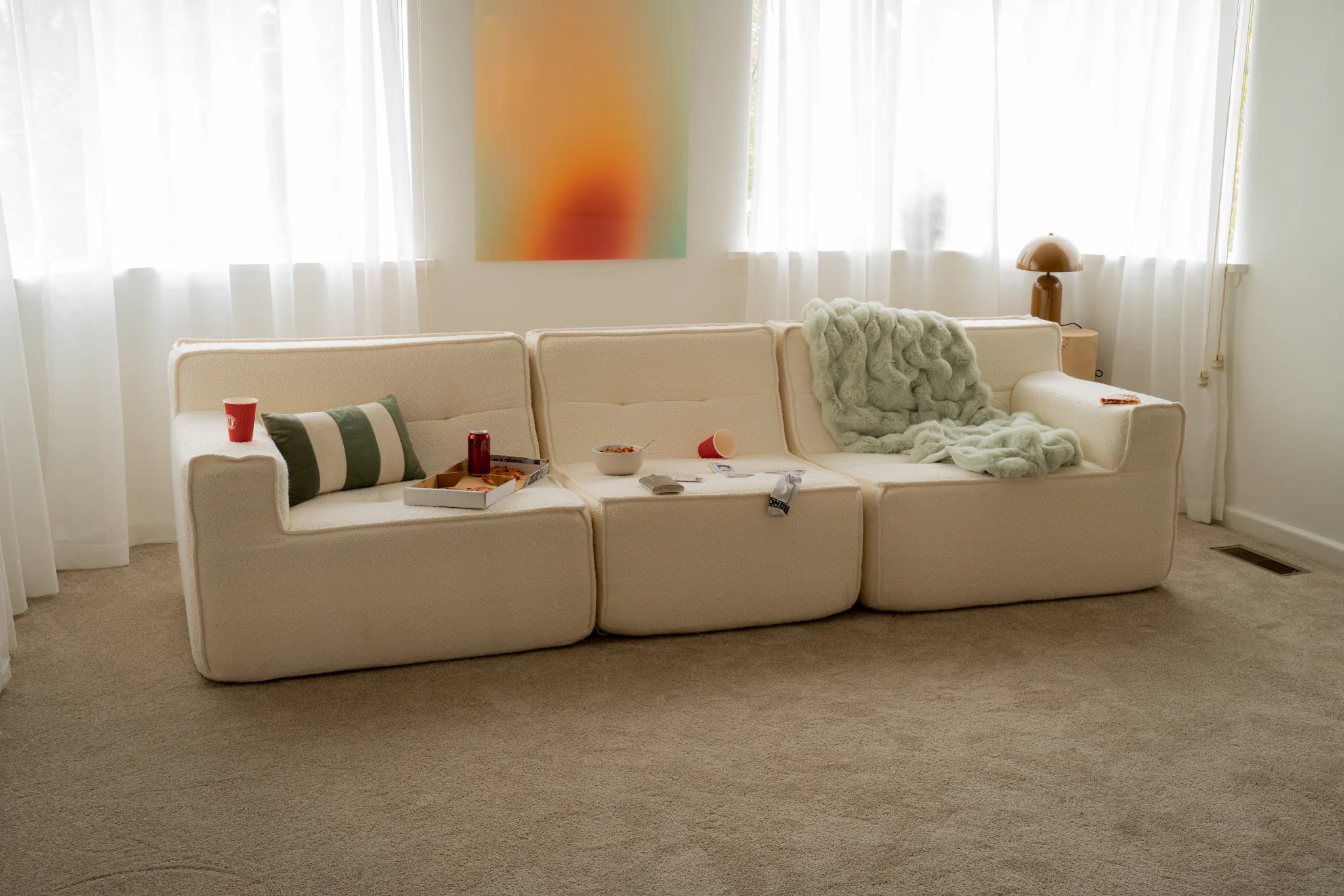 Beige sectional sofa with cushions and a small table in a room with white curtains and a colorful abstract painting.