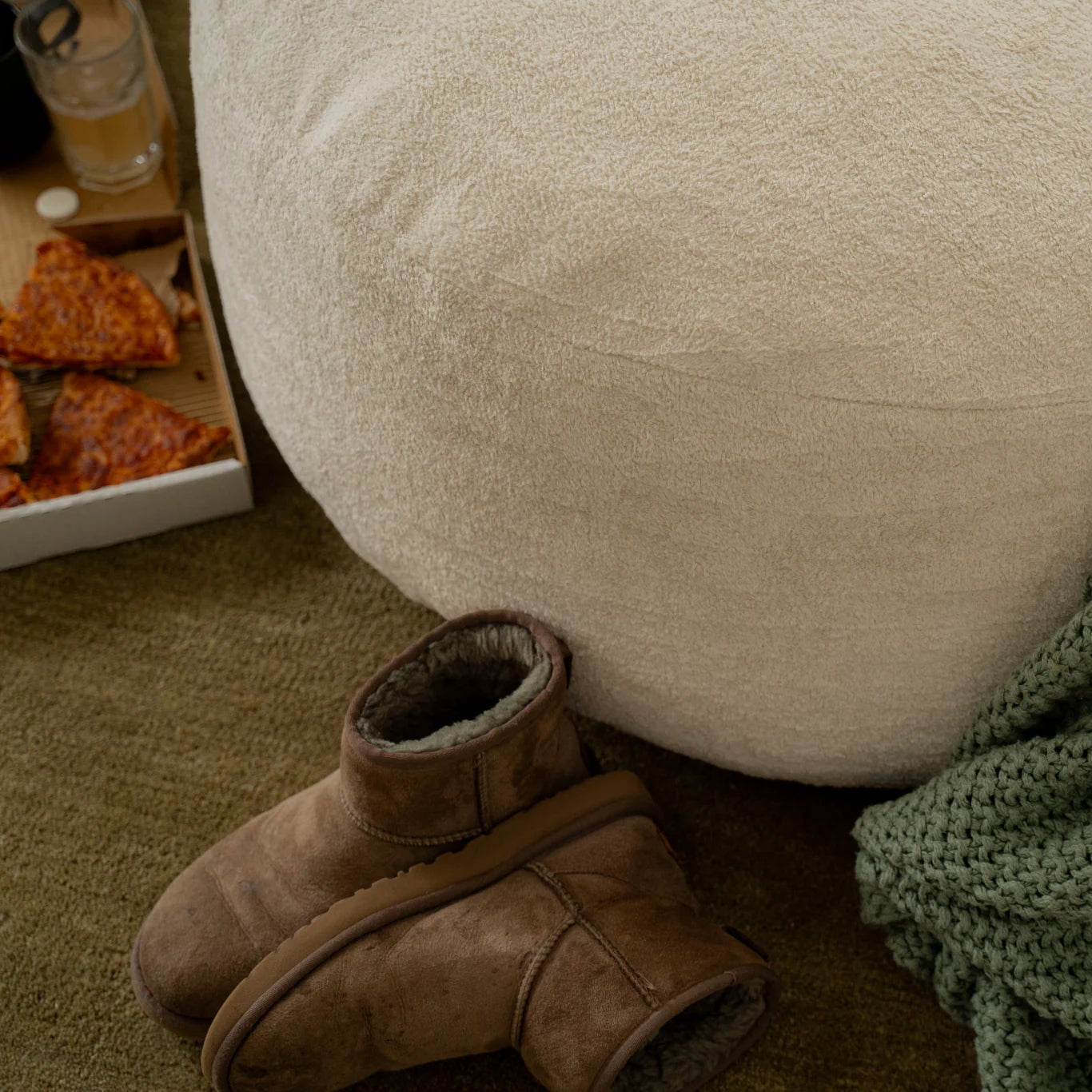 Brown boots next to a large white bean bag chair with a green blanket draped over it on a carpeted floor.