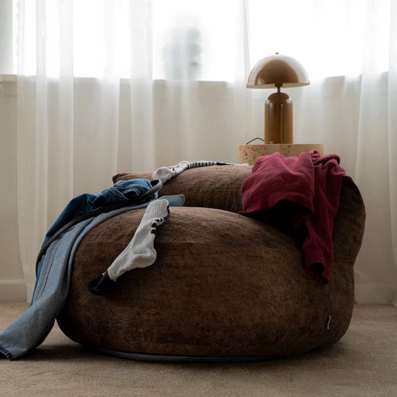 Brown bean bag chair with clothes on it in a room with white curtains and a lamp.