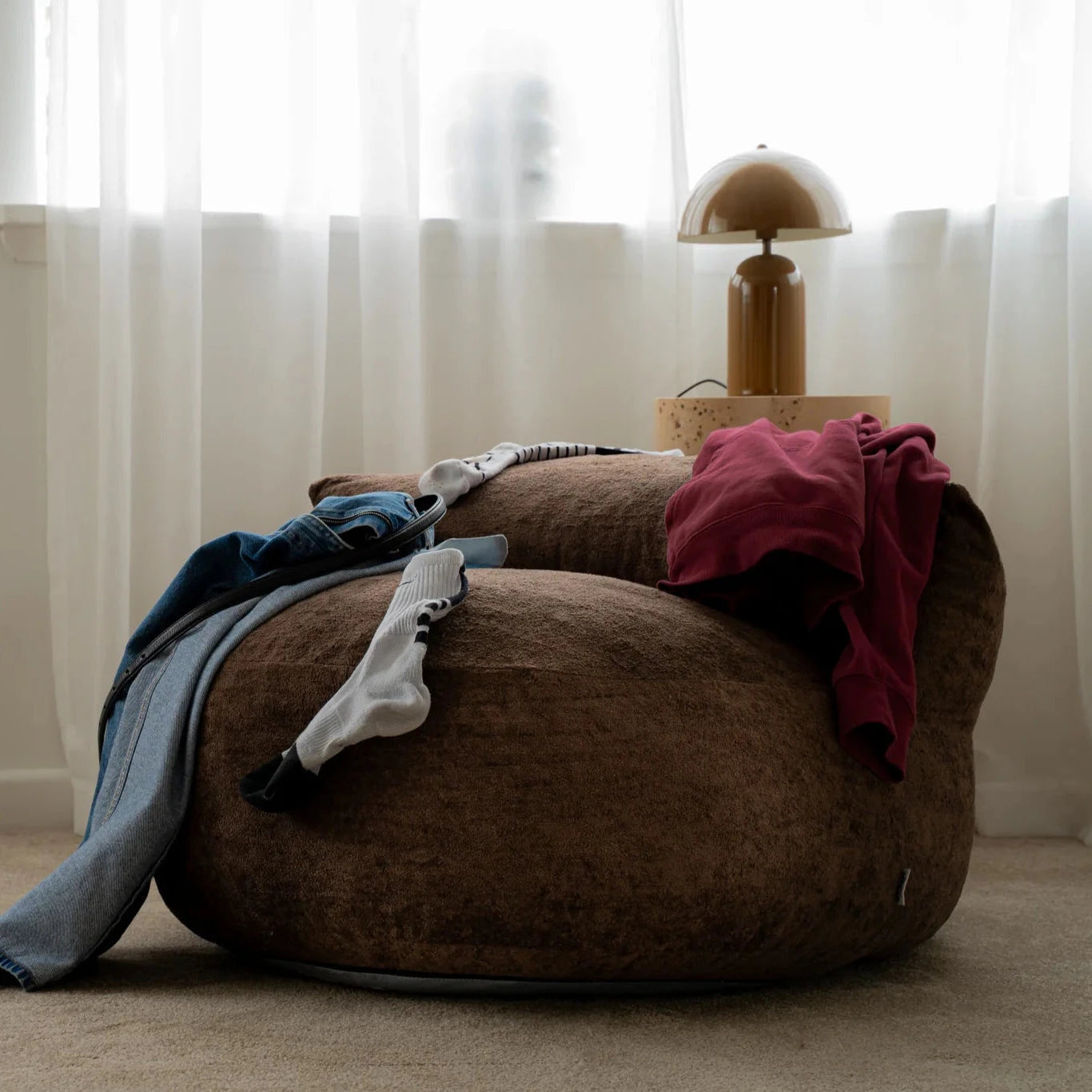 Brown bean bag chair with clothes on it in a room with white curtains and a lamp.