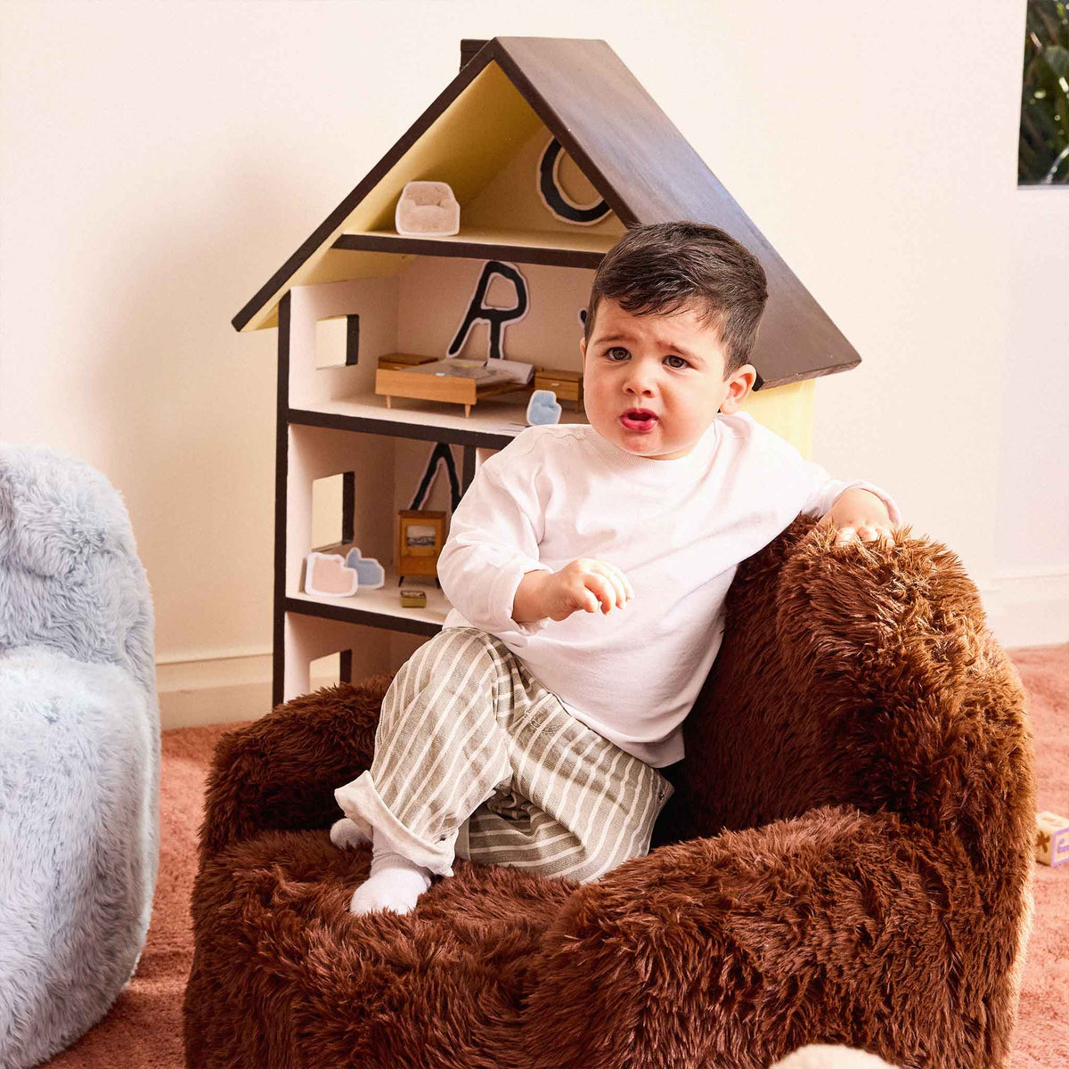 Toddler sitting in the brown Beari Kids Armchair beside a wooden dollhouse