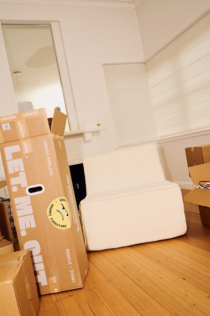 Unpacked cardboard box labeled 'Let Me Curl' on a wooden floor with a white couch in the background. 