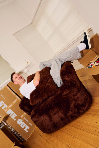 Person sitting on a large brown bean bag chair in a room with boxes and a light fixture. 