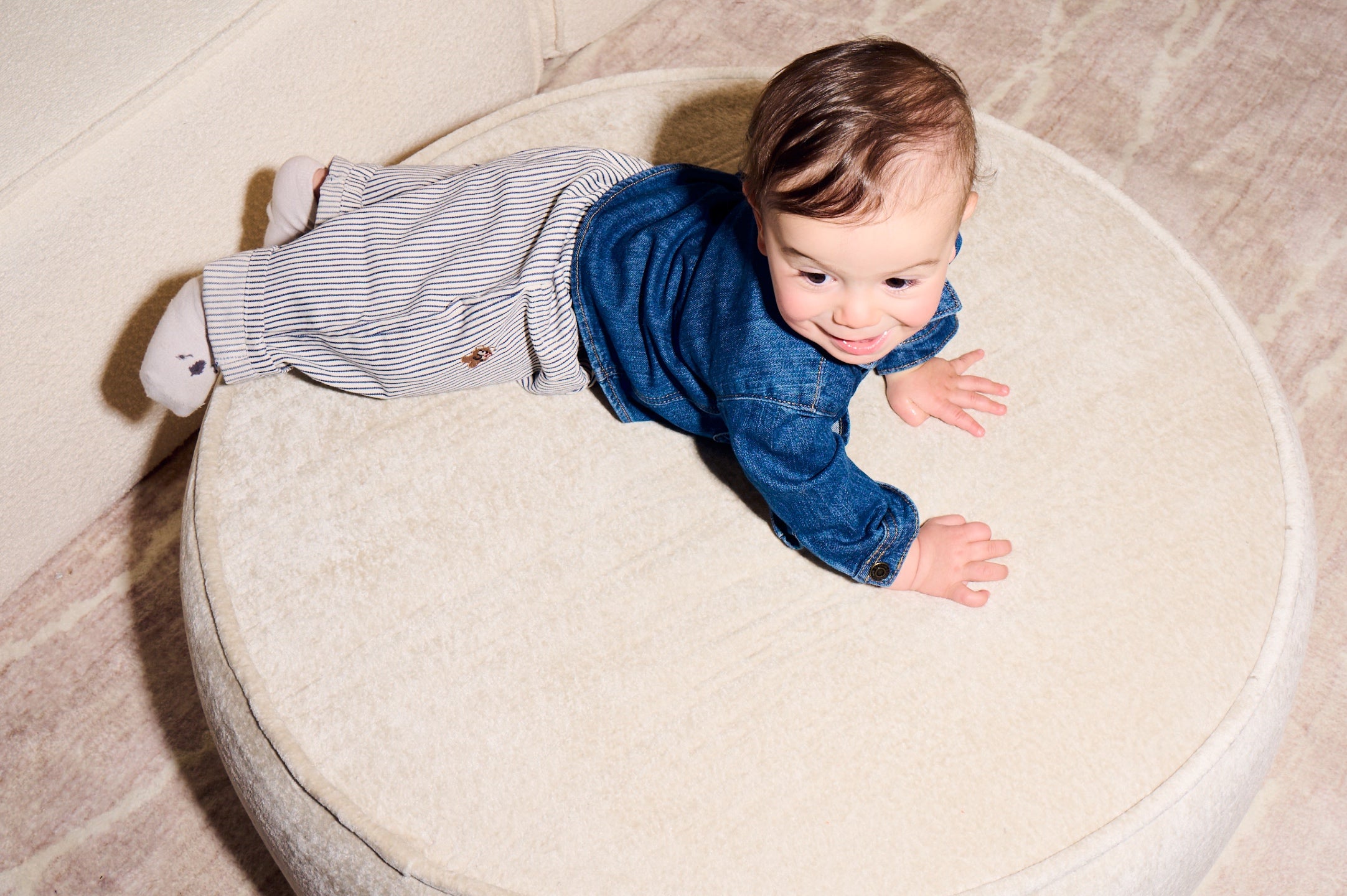 Baby in a blue shirt and striped pants crawling on a round, light-colored cushion. 