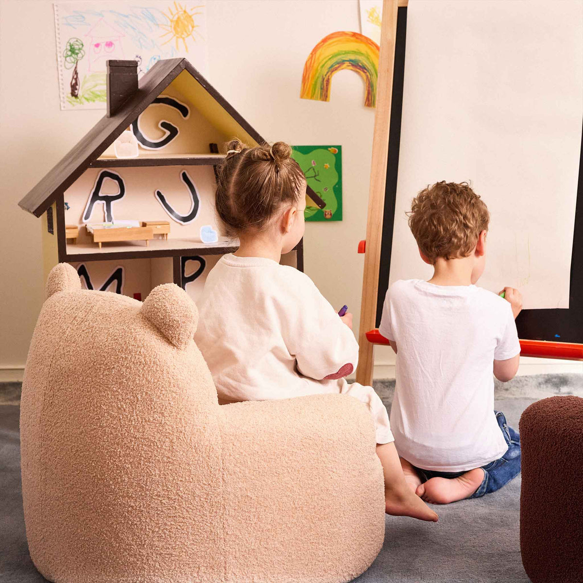 Two children drawing at an easel beside the cream Teddi Kids Armchair and a dollhouse 